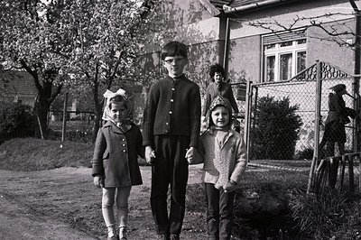 Three children stand outdoors, holding hands. The boy in the center wears glasses and a textured sweater, while the girls are...