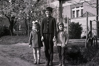 Three children stand hand-in-hand on a residential street lined with blooming trees. The central child, a boy, wears dark tro...