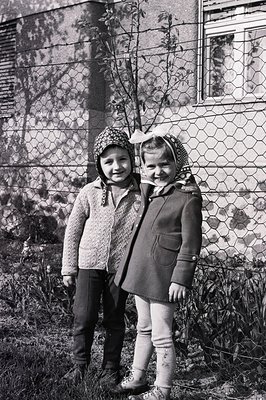 Two young children stand close together in a casual portrait. Both wear dark tights, sturdy boots, and headscarves. The boy w...