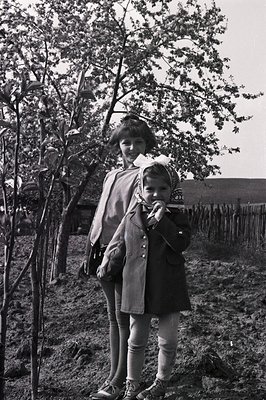 A young woman and child stand in a rural landscape, framed by blossoming branches. The woman wears a dark, wide-collar coat &...