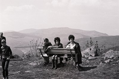 Three young adults seated on a wooden bench overlook a vast, hilly landscape. The individuals are dressed in 1970s-era attire...