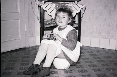 A young child sits playfully near a vintage wooden chair, patterned tiled floor visible. The room’s wallpaper is textured. Li...