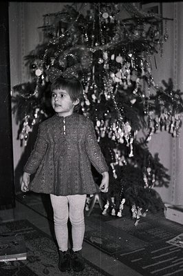 A young girl stands before a fully decorated Christmas tree. She wears a textured, A-line dress, tights, and dark shoes. Like...