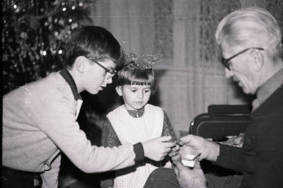 A boy and girl receive jewelry from an elderly man in a home setting. The boy, wearing glasses, helps secure a necklace on th...