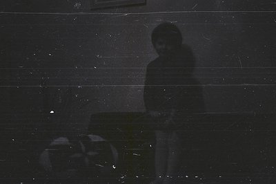 A grainy, high-contrast black and white image shows a young boy cast in shadow, seated on what appears to be a stool. The sce...