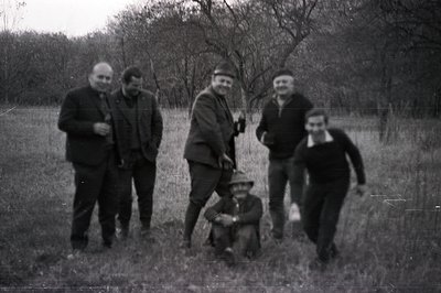Group portrait in a field. Four men stand or run, one sits, amidst tall grass & bare trees. Men wear suits, sweaters, and a f...