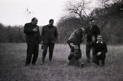 Four men in a grassy, wooded area. Two stand, two kneel and crouch, seemingly observing something off-camera. Men wear suits,...