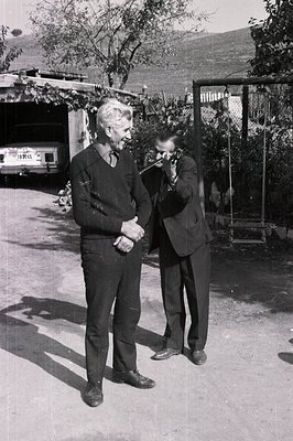 Two men stand outdoors. One in casual trousers and sweater gestures while the other, formally dressed in a suit, examines som...