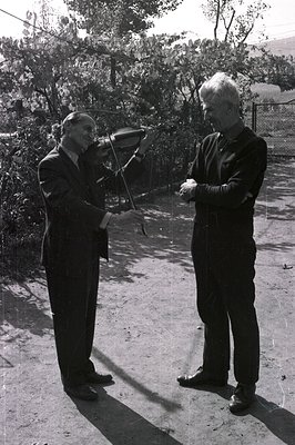 Two men stand outdoors on a gravel surface. One, formally dressed in a suit, appears to be presenting a violin to the other, ...