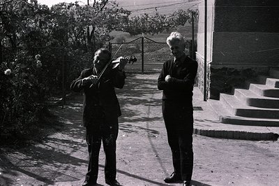 Two men are posed outdoors: one playing a violin, the other with arms crossed. An ornate gate and stone steps lead to a build...