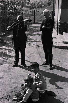 A seated boy with a bowl, likely soliciting money, is positioned before two men: one playing a violin, the other standing wit...