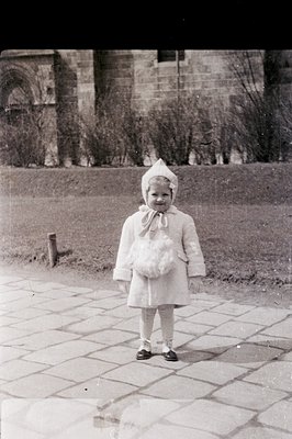 A young child, possibly a toddler, stands facing the camera, bundled in a winter coat and bonnet. The image exhibits vintage ...