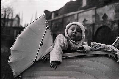 A toddler in a hooded sweater and bonnet gazes directly at the camera from a vehicle, likely a stroller. A partially opened u...