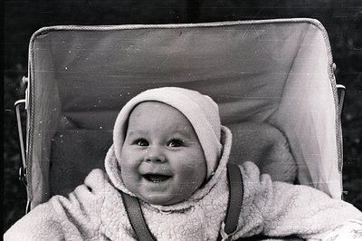 Infant smiles directly at camera, bundled in a textured, hooded garment and overalls while seated in a vintage stroller. The ...