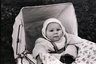 Baby sits securely in a vintage, padded pram featuring a rigid, peaked hood. Wearing a bonnet and appearing alert, the child ...