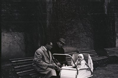 A candid, monochrome image features a man in a suit seated next to another man wearing a hat, with a baby in a vintage pram b...