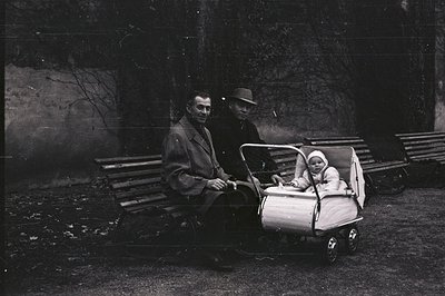 Two men and an infant are seated on a bench, an unusual stroller prominently displayed. Likely a candid portrait, the men are...
