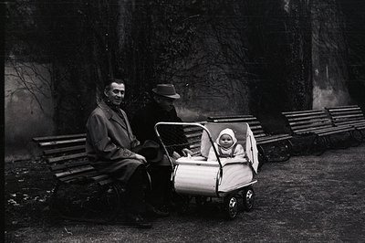 A seated man in a coat and a man in a hat observe a baby in a vintage, silver-toned pram in an outdoor setting. Wooden benche...