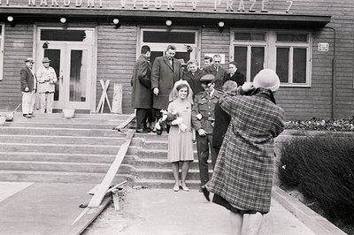 A young woman in a vintage dress & veil poses with a military officer outside a building in Prague. A photographer captures t...