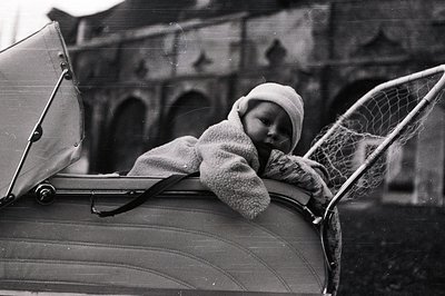 A young child, bundled in a textured sweater and cap, peers out from a vintage pram. The stroller's silver frame and canvas h...