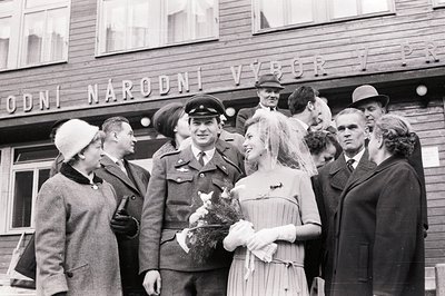 A newlywed couple poses with family and a military officer in front of a building, "Národní Výbor." The bride wears a veil an...