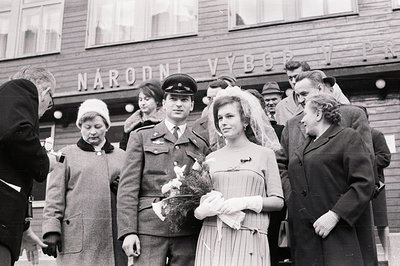 Formal portrait of a bride in a veil and gown, standing with a uniformed soldier and onlookers. The backdrop displays Cyrilli...