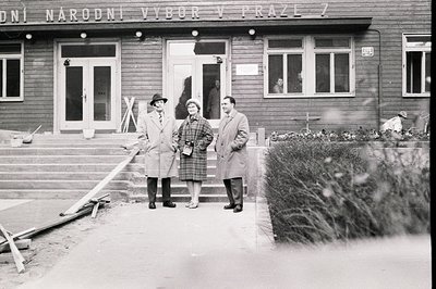 Three formally dressed people stand outside a building identified by signage as "Národní Výbor v Praze 7." The architecture i...