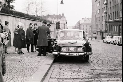A black and white street scene depicting a group of people gathered near a dark-colored DS Citroën sedan. The vehicle, parked...