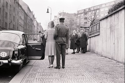 A military officer escorts a group of elegantly dressed women to a vintage car. The scene appears to be a formal departure, p...