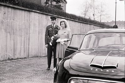 A formally attired groom in military uniform stands beside a bride with a veil and bouquet. They pose near a distinctive Tatr...