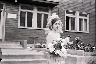 A young woman in a wedding veil and dress stands on concrete steps. She holds a bouquet wrapped in white fabric. Two men in h...