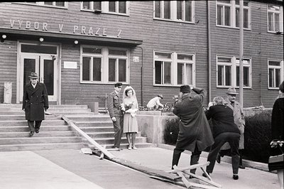 Exterior view of an apartment building ("Výbor v Praze 7") featuring a young couple in wedding attire, likely posed for a por...