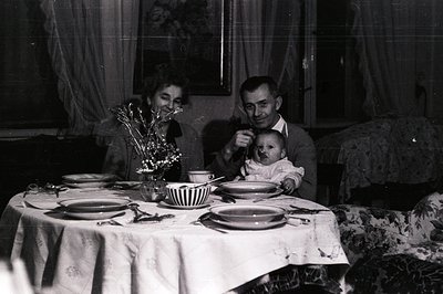 A family portrait featuring a couple and an infant, seated around a formally set table draped with a patterned cloth. Likely ...