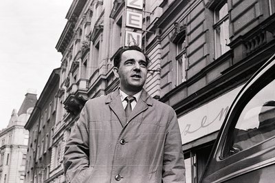 A man in a double-breasted coat and tie stands outside a building with ornate architecture. Likely 1960s street photography; ...
