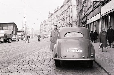 A man in a long coat stands beside a vintage sedan on a cobblestone street lined with early 20th-century buildings. Pedestria...