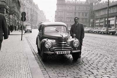 A man stands proudly beside a Saab 93, a classic early model, on a cobblestone street. Mid-century urban scene shows other ve...