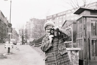A man, wearing a checked coat and hat, operates a vintage camera, likely a Rolleiflex, in an urban setting. The architecture ...