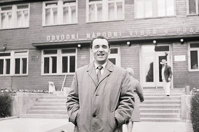 Man in a double-breasted coat smiles at the camera, positioned outside a building identified as “Obvodní Národní Výbor v Praz...
