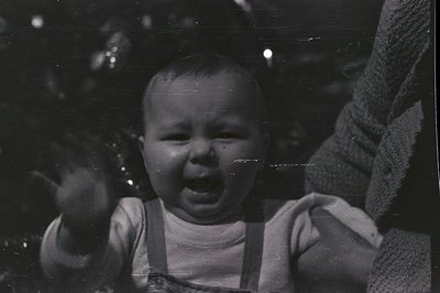 Infant in overalls, displaying a distressed expression, held by an unseen figure. Grainy black and white photograph, likely a...