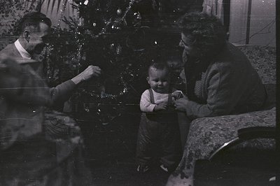 A young child in overalls stands between a seated man and woman near a decorated Christmas tree. The family appears to be cel...