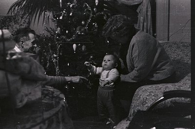 A young child stands near a decorated Christmas tree, reaching toward a seated man in a suit. A woman with curly hair sits ne...