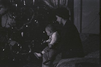 A seated woman holds a baby, posed near a Christmas tree. Interior scene; lighting is subdued. Likely a family portrait, 1920...