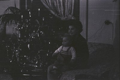 A young child in overalls sits on a woman’s lap, both positioned near a brightly lit Christmas tree. The image, captured in b...