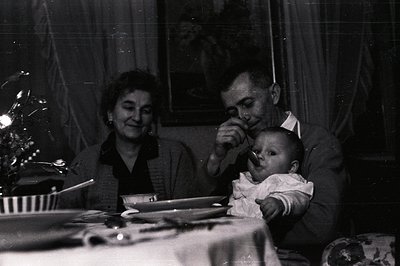 A tender moment: a man cradles a baby while a woman watches, seated at a table. Traditional dress and the visible table setti...