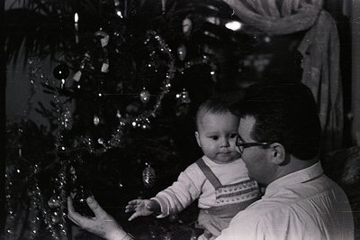 A father, wearing glasses and a collared shirt, holds a baby reaching toward a decorated Christmas tree. Classic vintage orna...