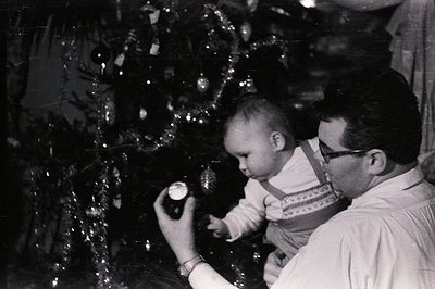 A man, wearing glasses and a collared shirt, holds an infant, likely captivated by an ornament on a heavily decorated Christm...