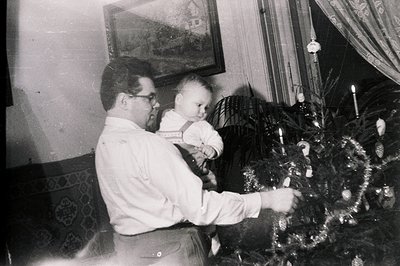 A man in a collared shirt holds an infant while decorating a Christmas tree with tinsel. A framed picture hangs on the wall, ...