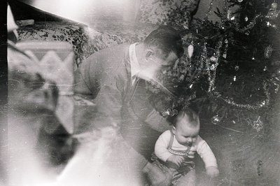 A father tenderly interacts with his young child near a decorated Christmas tree. Interior scene shows patterned wallpaper an...