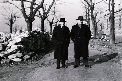 Two formally dressed men in overcoats and hats stand facing the camera on a gravel path. A pile of rubble and leafless trees ...