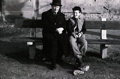 A stern-faced elder and a young boy sit side-by-side on a weathered wooden bench. Both wear dark hats and heavy coats, sugges...